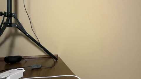 A video of a corner in a room with beige walls. A black tripod and cables are on a wooden floor, and a white remote control is visible. The scene is minimalist and utilitarian.