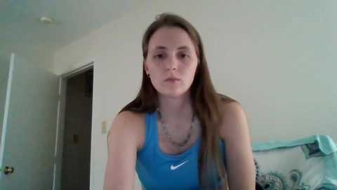 A video of a young Caucasian woman with long brown hair in a blue Nike tank top, standing indoors with a white wall and open door in the background.
