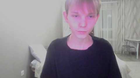 A video of a young boy with short, light brown hair wearing a black shirt, standing indoors against a gray wall. Background includes a radiator, curtains, and a white chair.