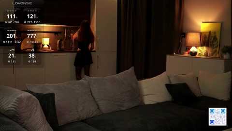Video of a modern living room at night with dim lighting. A woman in a black dress stands by a kitchen counter, while a grey sectional sofa with dark pillows is visible in the foreground.