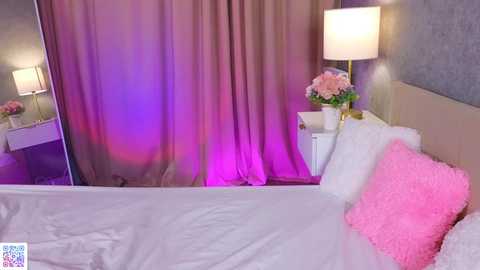 Video of a modern bedroom with a white bed, pink fluffy pillow, mauve curtains, a mirrored wall, and a white nightstand with a vase of flowers and a lamp.