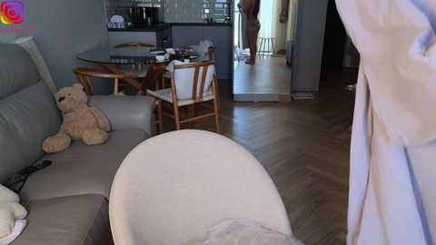 Video of a modern living room with a beige sectional sofa, wooden dining table, and a teddy bear, leading to a kitchen and hallway.