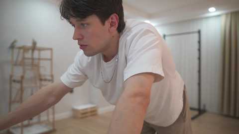 Video of a young, light-skinned man with short brown hair, wearing a white t-shirt and beige pants, leaning forward, in a minimalist, white-walled room with beige flooring, ladder, and empty boxes.