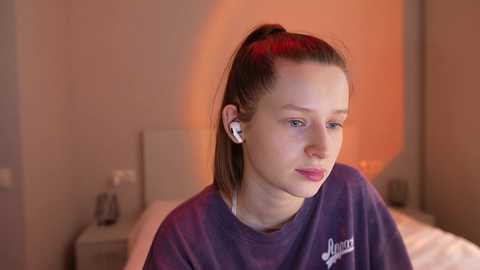 Video of a young woman with fair skin and straight brown hair tied back, wearing a dark purple shirt and white earbuds, in a softly lit, beige-colored bedroom.