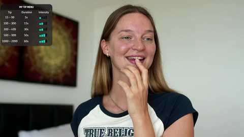 Video of a smiling young woman with long brown hair, wearing a white and navy T-shirt, holding a finger to her lips in a bedroom with a black headboard and abstract art on the wall.