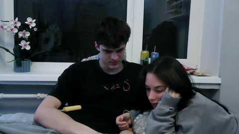 Video of a young man and woman in a hospital room. He has short dark hair, wears a black t-shirt, and holds a wooden stick. She has long dark hair, wears a grey hoodie, and rests her head on his shoulder. A white window with a pink orchid in a vase is behind them.