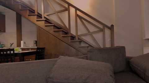 Video of a modern, minimalist living room with a gray sectional sofa and wooden staircase with glass railings leading to an open kitchen area with white countertops and wooden cabinets.