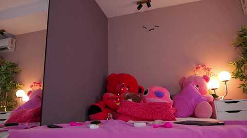 A video of a pink-themed bedroom with a large teddy bear, plush toys, and a mirror reflecting the room's decor, featuring a white dresser with lamps, potted plants, and an air conditioner.