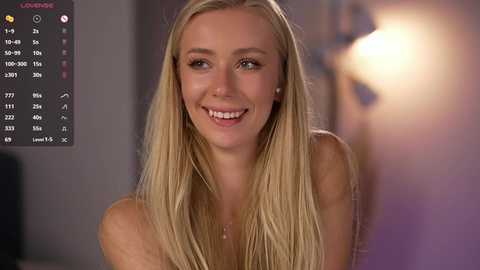 Video of a smiling, blonde woman with straight hair, wearing a strapless top, in a softly lit room with a blurred, warm-toned background.