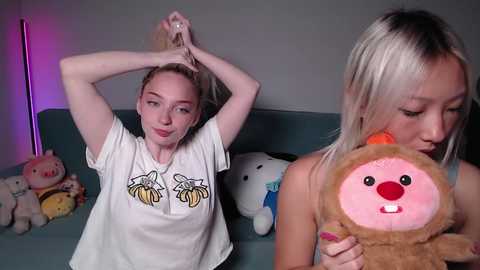 Video of a Caucasian woman with long blonde hair, wearing a white t-shirt with banana graphic, playfully holding her hair, standing next to a topless Asian woman holding a plush toy in a dimly lit room.