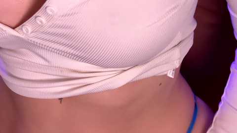 Close-up video of a woman's ribbed, white, button-up top, partially unbuttoned, revealing a blue thong, highlighting her midriff and cleavage, against a blurred dark background.