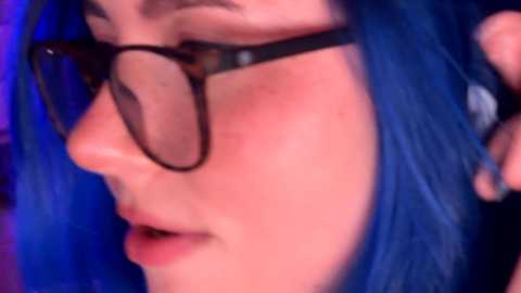 Video of a close-up of a woman's face with blue hair, wearing glasses, and a black top, blushing with a hint of red on her cheeks, set against a blurred, dark background.