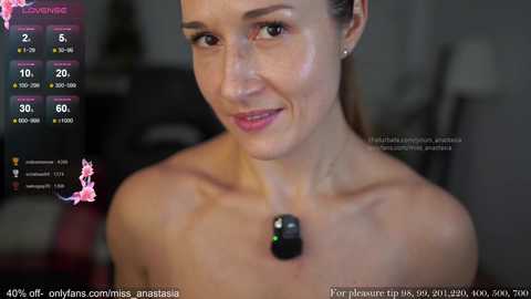 Video of a smiling, topless woman with fair skin and brown hair, wearing a black wireless headset, in a dimly lit room with a blurred background.