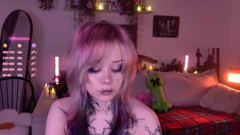 Video of a young woman with pale skin and long, pastel-colored hair, covered in black ink tattoos, sitting on a bed adorned with plush toys, surrounded by dimly lit, cozy bedroom decor.