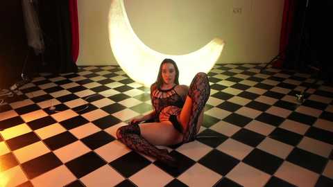 Video of a woman with long hair, wearing black lingerie and thigh-high boots, seated on a black-and-white checkered floor, in a room with a glowing crescent moon backdrop.