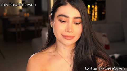 Video of a young woman with long black hair, fair skin, and light makeup. She is indoors, wearing a strapless top, with a cozy, dimly lit background featuring a fireplace and blurred furniture.