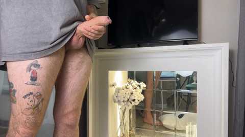 Video of a man with a tattooed leg, holding an erect penis, standing in front of a white-framed mirror reflecting a living room with a flat-screen TV, a white vase, and a chair.