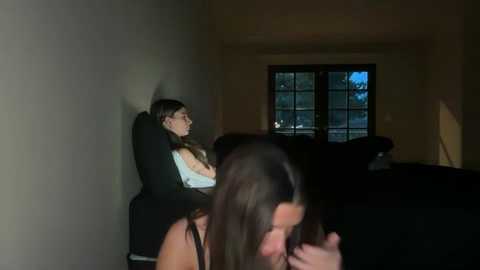 Media: Video of a dimly lit living room with a boy sleeping on a couch and a woman in the foreground, blurry, sitting. A window with a dark sky view is visible.