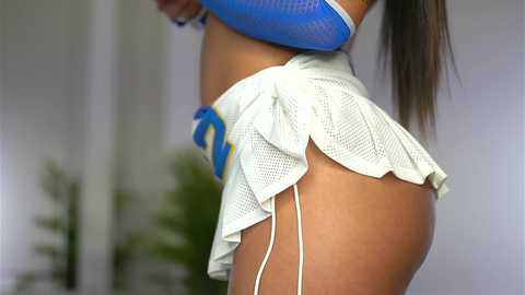Video of a woman in a blue mesh crop top and a white mesh skirt with a blue and yellow trim, highlighting her toned, tanned thighs and buttocks.