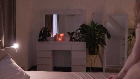 Video of a dimly lit bedroom with a white dresser, a potted plant, candles, and a large mirror; a person's arm is visible in the foreground.