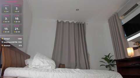 Video of a minimalist bedroom with a neatly made bed, beige curtains, a wooden dresser, a TV on the wall, and a potted plant.