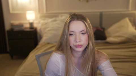 Video of a young woman with long, straight blonde hair, wearing a white off-shoulder top, seated on a bed in a softly lit, cozy bedroom with beige bedding and a nightstand with a lamp.