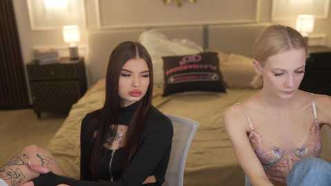 Video of two women in a bedroom; one with long dark hair, black top, tattoos, and red lipstick, the other with blonde hair, floral top, and neutral expression.