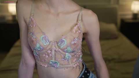 Video of a slender, light-skinned woman with small breasts, wearing a sheer, floral lace bralette with pastel colors and a black leather skirt. Background shows a dimly lit bedroom with a bed and nightstands.