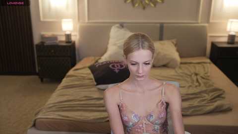 Video of a blonde woman with fair skin wearing a floral-patterned sheer dress, sitting on a bed in a dimly lit, beige room with two nightstands, a lamp, and a decorative gold star.
