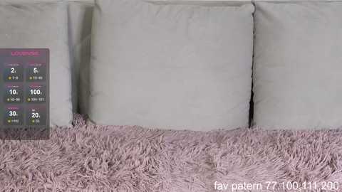 Video of a modern living room featuring a plush, soft, light pink shag rug. The background consists of a white sofa with three large, rectangular, white pillows. A digital screen displaying a \"Lavacore\" logo and temperature readings is placed on the left side.