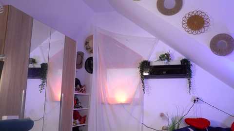 Video of a cozy, modern attic bedroom with a white ceiling, white walls, and a hanging white canopy bed. The room features a wooden wardrobe, potted plants, and decorative circular wall hangings. Warm lighting creates a serene atmosphere.