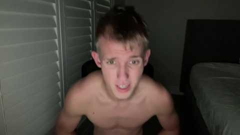 Video of a young, shirtless white male with fair skin, short brown hair, and a startled expression, leaning forward against white blinds in a dimly lit bedroom.