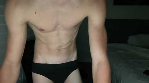 Video of a shirtless, slender, fair-skinned man in black briefs, with visible ribs and a toned abdomen, standing in a dimly lit bedroom with white sheets and a dark headboard.