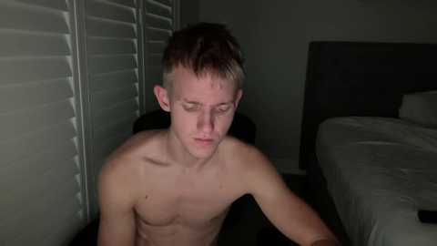 Video of a shirtless, fair-skinned young man with short, light brown hair and a lean physique, sitting on a bed in a dimly lit room with white blinds and a gray headboard.