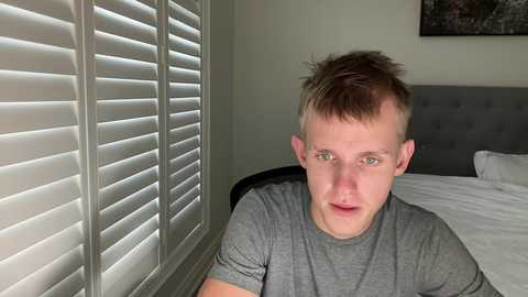 Video of a fair-skinned, short-haired, young man in a gray t-shirt, sitting on a bed with white linens, in a room with white shutters and a framed photo on the wall.