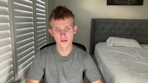 Video of a young Caucasian man with short brown hair, wearing a gray t-shirt, sitting on a chair in a modern, minimalist bedroom with white blinds, a gray upholstered headboard, and neatly made bed.