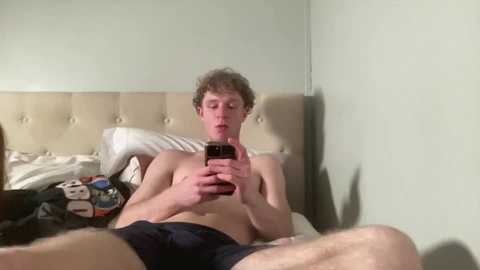 Media: Video of a shirtless, fair-skinned, curly-haired young man in black shorts, sitting on a bed with beige headboard, holding a smartphone, surrounded by scattered toys and pillows.
