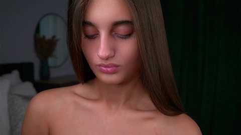 Video of a young, light-skinned woman with long brown hair, closed eyes, and a serene expression, wearing pink lipstick. Background shows a dark curtain and a white pillow.