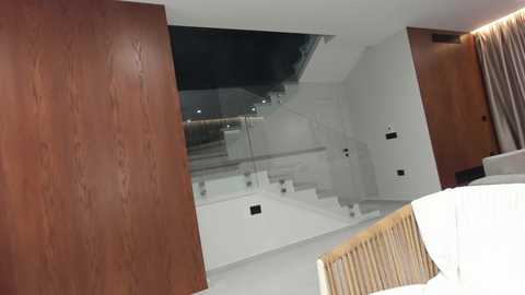 Video of a modern bedroom with a wooden panel wall on the left, glass staircase in the center, and a wooden chair with white pillows in the foreground.