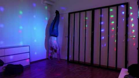 Video of a person doing a handstand against a white wall with colorful string lights, in a room with wooden floor and white dresser.