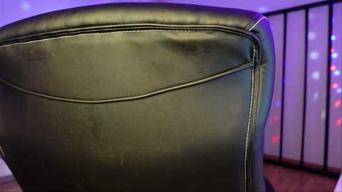 Video of a black leather chair with a slightly worn texture, featuring a slight crease on the left side. Background shows a purple light, wooden floor, and a black metal railing.