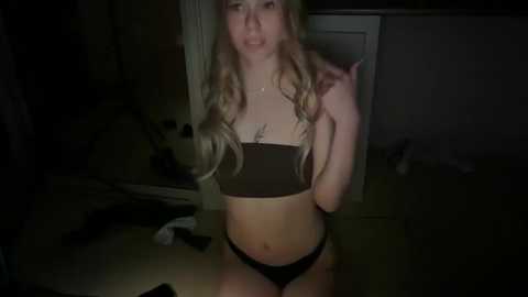 Video of a young blonde woman with wavy hair, wearing a black bandeau top and black panties, posing indoors in a dimly lit room with scattered clothes and a white door.