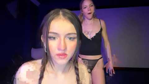 Video of two women: a young, pale-skinned brunette in a sequined top, and a blonde in a black lace bra and panties, standing in a dimly lit room with purple lighting.
