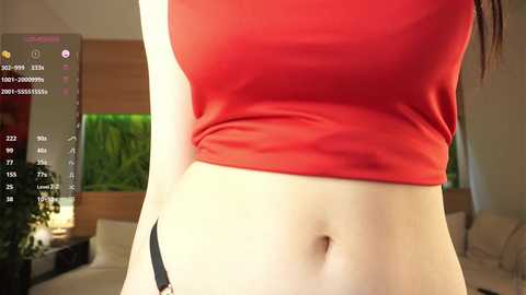 Video of a light-skinned woman in a red crop top, highlighting her flat stomach and toned abs. The background shows a modern living room with a green aquarium and plants.