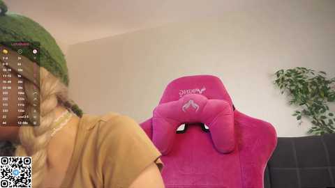 A video of a blonde woman in a green hat, seated on a pink \"Shinobi\" gaming chair, with a green plant and a gray couch in the background.