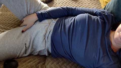 Video of a person wearing a blue long-sleeve shirt and gray sweatpants, lying on a quilted brown bedspread, with a hand resting on their abdomen.