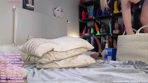 A video of a messy bedroom with a beige quilted blanket, a stuffed animal, and a wicker basket. The background features colorful sex toys on a bookshelf and a woman in a black top partially visible.