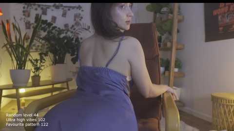 Video of a woman with dark hair, wearing a blue dress, seated in a brown chair in a dimly lit room with potted plants and wall decor.