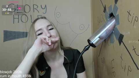 Video of a blonde woman with light skin, wearing a black shirt, making a peace sign with her fingers. Background features a hole with a metal nozzle and \"GLORY HOLE\" written on a beige wall.