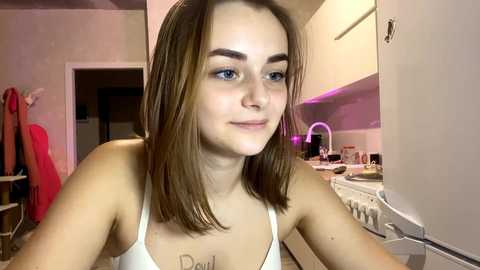 Video of a young woman with fair skin and straight, light brown hair, wearing a white spaghetti-strap tank top. She has a tattoo under her collarbone. The background shows a cluttered kitchen with a sink and cabinets.